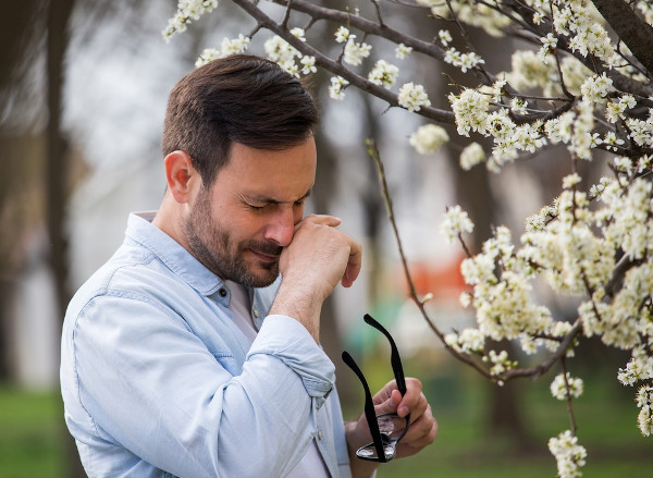 Homem perto de árvore florida coçando o nariz, demonstrando ser alérgico a pólen.