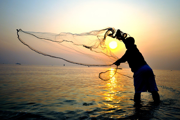 Pessoa lançando uma rede de pesca no mar com o nascer do Sol ao fundo.