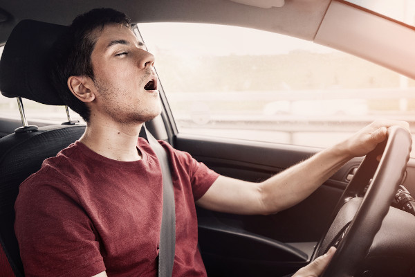 Homem dormindo com a boca aberta segurando o volante, dentro de um carro; narcolepsia pode provocar acidentes de trânsito.