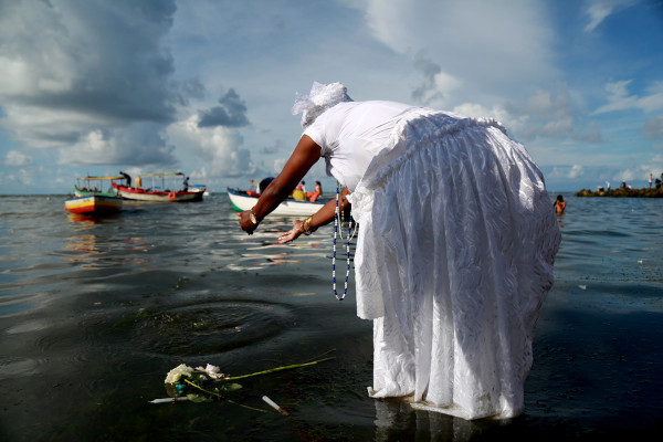 Mulher com vestes do candomblé lança flores nas águas do mar no Dia de Iemanjá; ao fundo, alguns barcos.