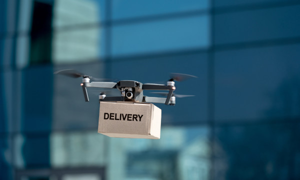 Drone carrega caixa, onde se lê: “Delivery”.