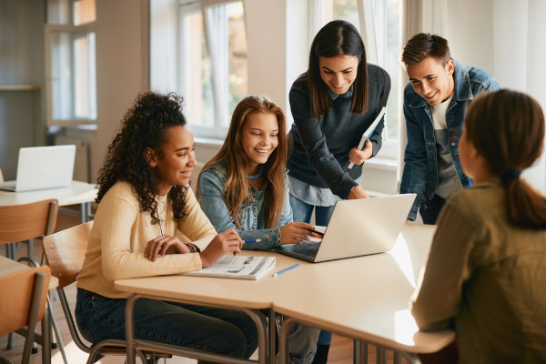 Grupo de jovens em uma sala de aula ao redor de um notebook, atividade comum durante cursos de férias.