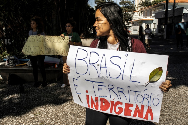 Mulher indígena segurando cartaz com escrito “Brasil é um território indígena” em defesa dos direitos dos povos originários.