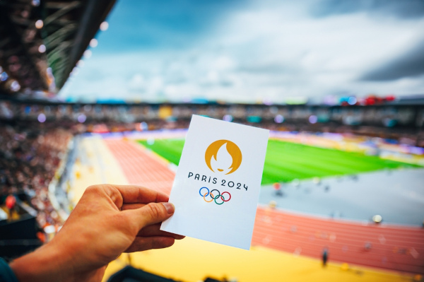 Pedaço de papel com o emblema das Olimpíadas de Paris 2024 aparece em primeiro plano, ao fundo um estádio olímpico. 