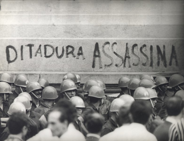 Frase “Ditadura assassina” gravada em um muro, um protesto pelo fim da Ditadura Militar; em frente, militares e civis. [imagem_principal]