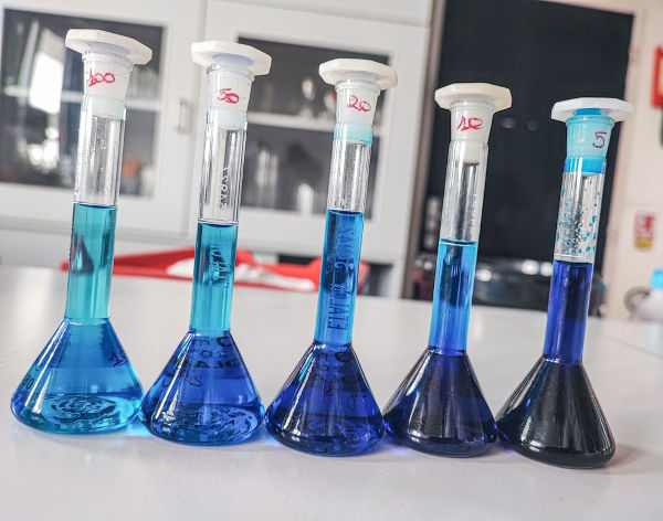 Cinco frascos com soluções de cor azul em um laboratório.