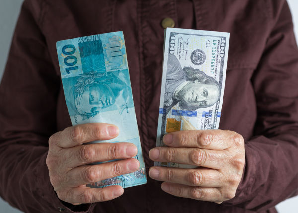 Mãos segurando cédulas de real e de dólar, em alusão ao câmbio financeiro. [imagem_principal]