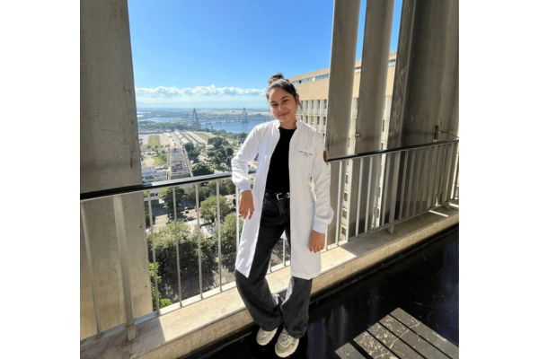 Mayumi, aprovada em medicina no Enem, de jaleco em uma janela com a vista da cidade no fundo.