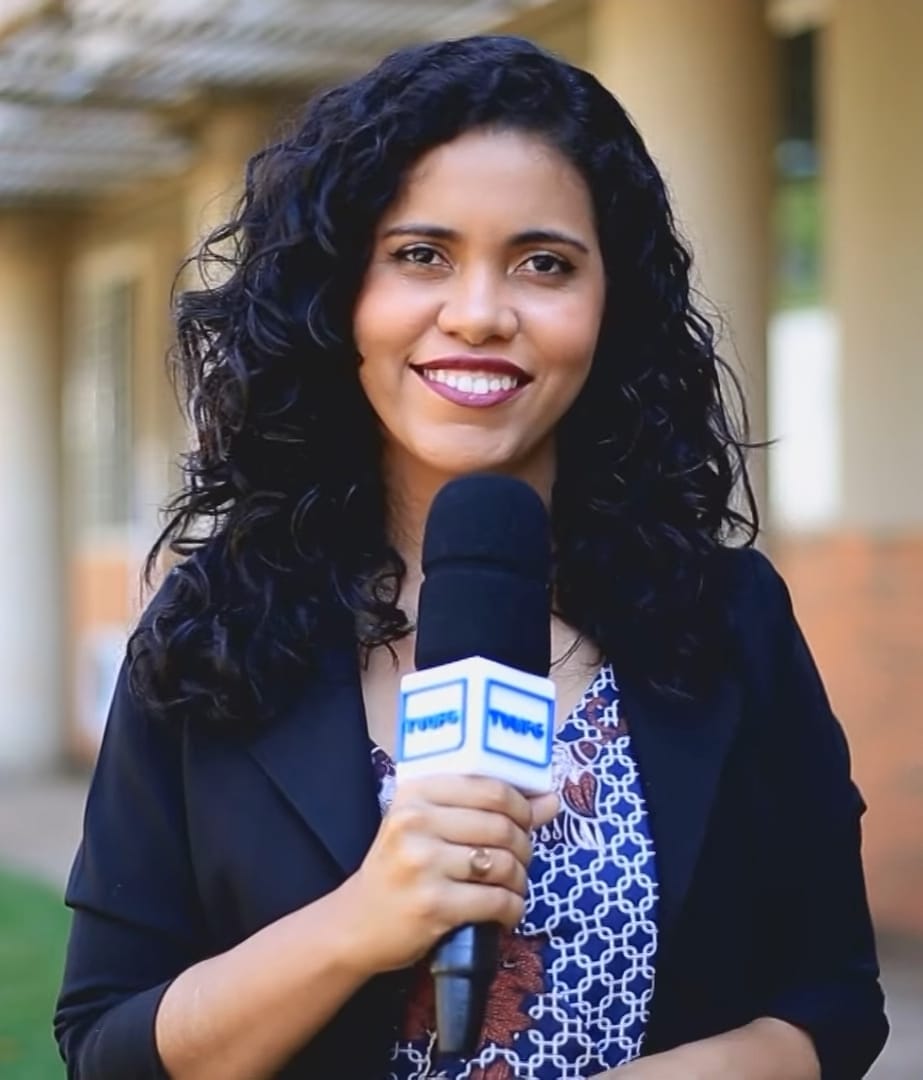 Anna Clelma é uma jornalista negra e está sorrindo e segurando um microfone